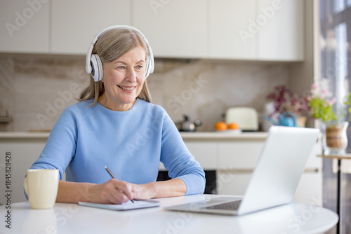Smiling senior woman wearing headphones and taking notes in a notebook while actively engaging in an online e-learning course on her laptop from a home kitchen setting