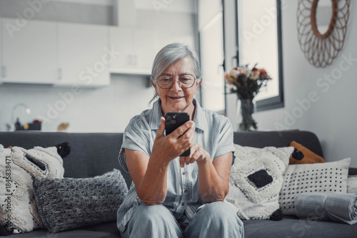 Elderly woman sits on a sofa at home using her smartphone to browse and shop online. She focuses on digital communication and enjoys a modern e-commerce lifestyle.