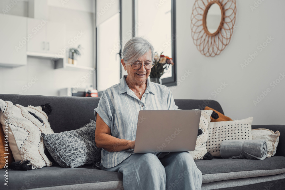 © Daniel - Elderly woman sits on a sofa at home using her computer to browse and shop online. She focuses on digital communication and enjoys a productive e-commerce routine.