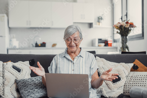 Elderly woman sits on a sofa at home smiling during a video call with family and friends. She enjoys connecting and sharing happy moments through modern technology.