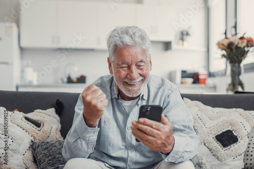 Elderly man celebrates at home while checking his smartphone. He feels joyful and euphoric after winning or succeeding online.