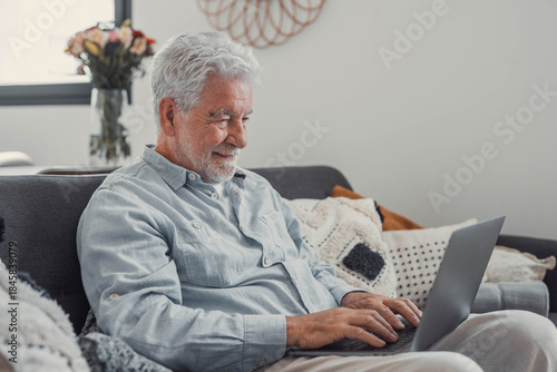 Elderly man sits on a sofa using his computer to browse and shop online. He enjoys a productive lifestyle while staying connected digitally at home.