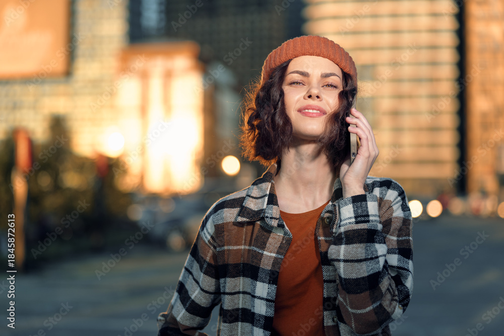 Fototapeta premium woman phone urban golden hour candid lifestyle beanie portrait: Young woman smiling on phone in city street at golden hour glow, casual fashion and curly hair, authenticity, mindful living and