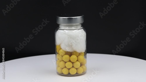 A close-up studio shot of a transparent glass vial containing numerous small, round yellow pills at the bottom and a layer of white cotton on top, capped with a silver lid. 