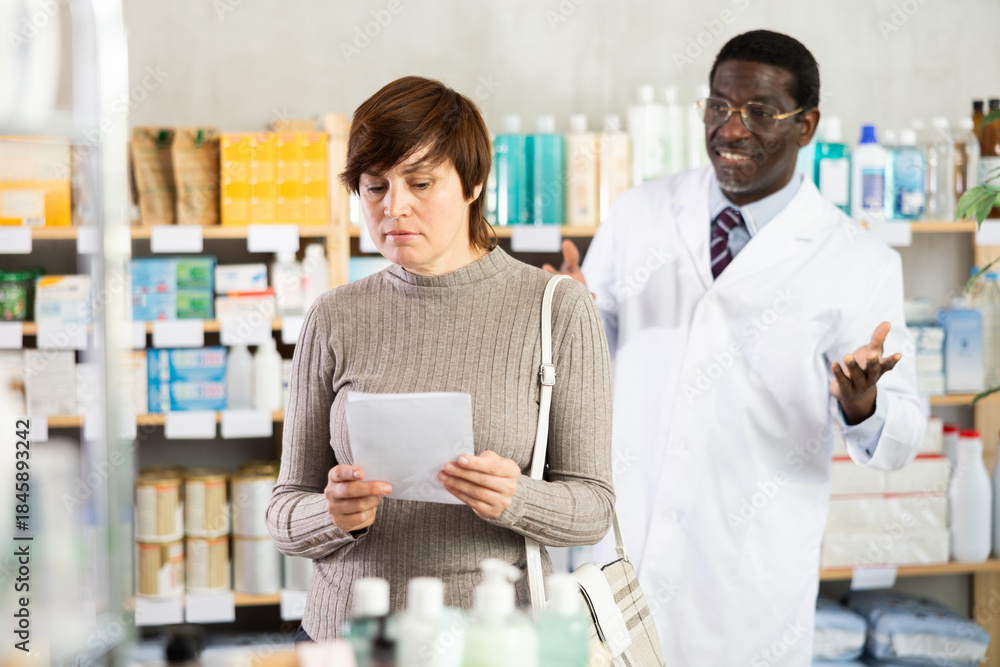 Obraz premium In sales area of drugstore, woman with prescription looks at display cases, trying to find multivitamins for adults in open display. African male pharmacist informs about possibility of replacing