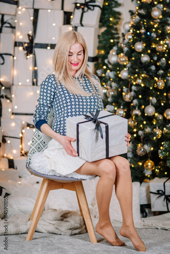Woman with long blonde hair sitting on chair, holding beautifully wrapped gift box, surrounded by festive Christmas decorations and sparkling lights