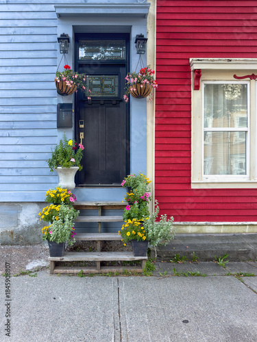 A blue house with a black door. There's a small window in the door, and over called a transom window. The adjoining house is vibrant red with white trim. The steps to the door have colorful flowerpots
