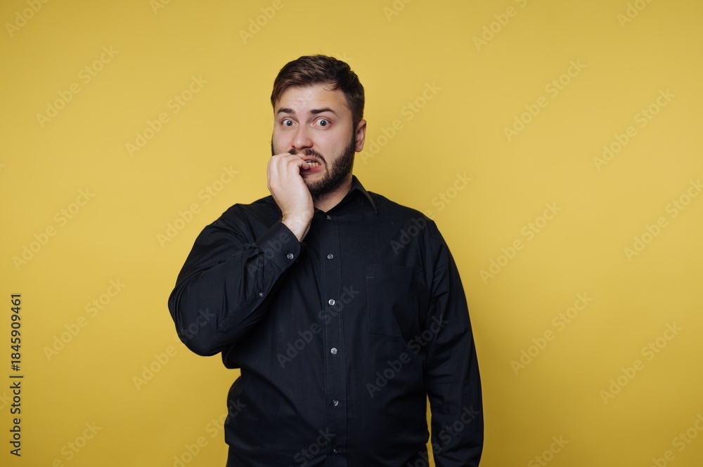 Fototapeta premium Man shows surprise and concern with hand on mouth in front of yellow background during daytime in indoor setting