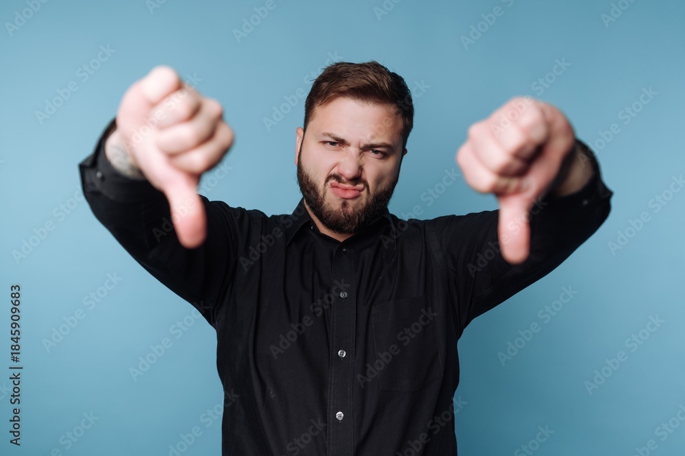 Fototapeta premium Man shows thumbs down gesture against blue background, expressing disapproval during a casual moment