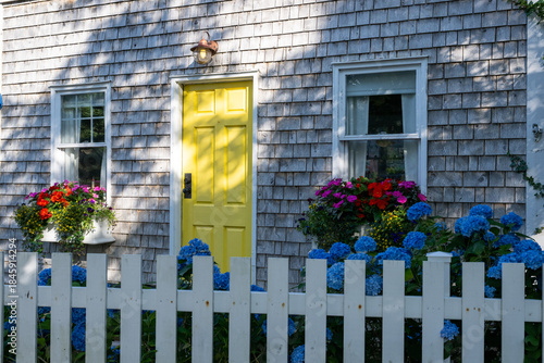 The exterior entrance to a small country cottage with grey cedar shakes, double hung windows, and a bright yellow solid panel door. There are colorful flowers, shrubs, and flower boxes in the yard.