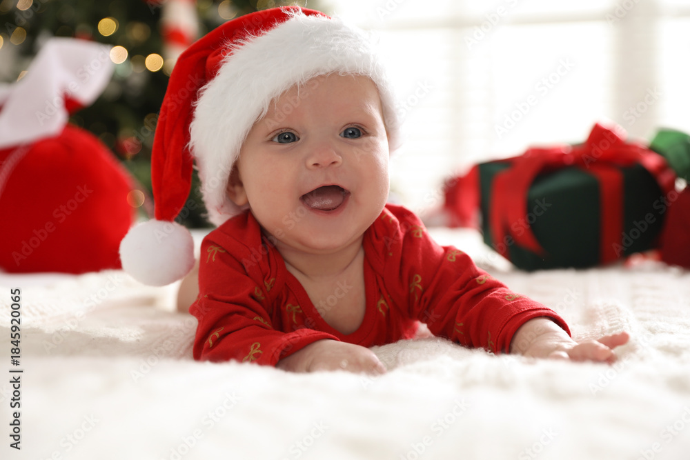 Obraz premium Cute baby in Santa hat on bed against blurred lights indoors. Christmas season