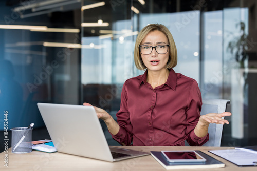 Billede på lærred Businesswoman sitting at her desk with a laptop, shrugging with open hands, conv