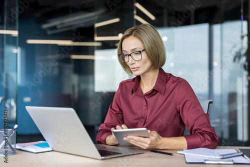 фотографія Confident businesswoman with eyeglasses working diligently on a laptop and holdi