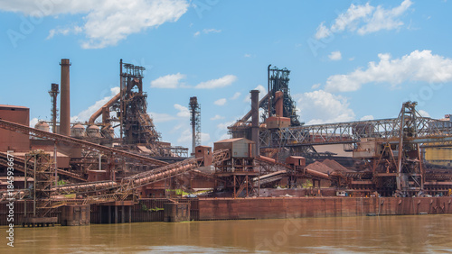 Industrial landscape of a massive, rust-colored mining complex by a muddy river under a blue sky with white clouds