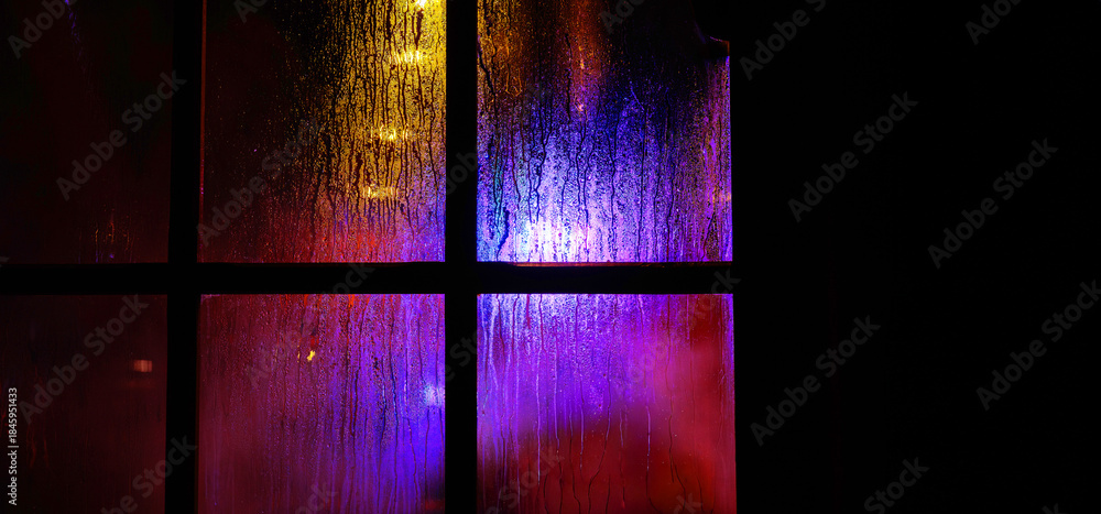 Fototapeta premium A dark window pane with rain droplets at night. Blurred yellow and blue festive christmas lights through the wet glass covered with condensate drops. Abstract background.