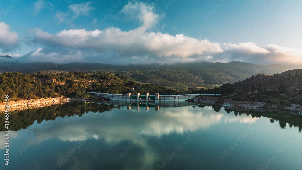 Fototapeta premium A serene reservoir and dam in South Spain, surrounded by lush hills and mountains, reflecting clouds and sky on calm water. Ideal for nature, travel, and landscape inspiration.
