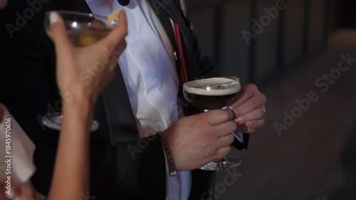 CloseUp Formal Guests Holding Martini Glasses At Elegant Gala Reception Businessman In Tuxedo Cradles Espresso Martini While Companion