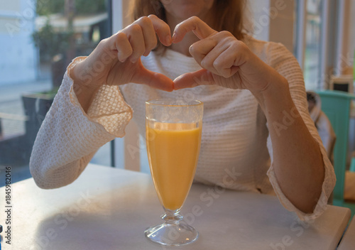 A woman forms a heart shape with her hands over a glass of fresh orange juice while sitting at a coffe table