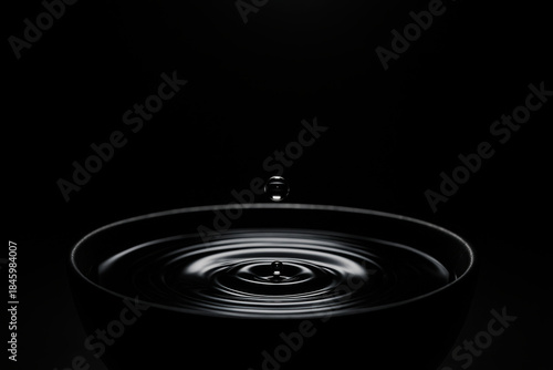 Single water droplet falling into dark reflective bowl creating circular ripples in minimal black background