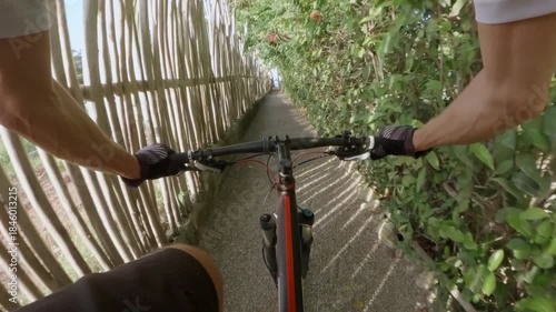 POV. Person rides a mountain bike along a narrow gravel path surrounded by green bushes and wooden fences goes to ocean.