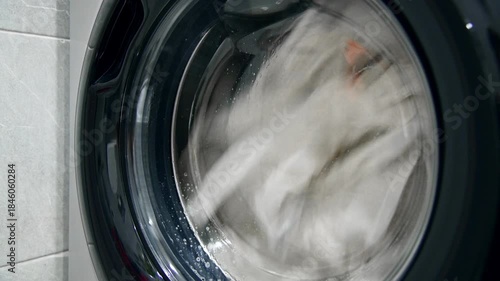 A close-up of a domestic washing machine window. White linens are washed, spinning and tumbling in the washing machine drum. Foam and bubbles run down the glass door viewing window.