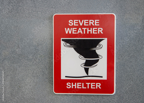 A red sign for a Severe Weather Shelter against a gray wall with a graphic of a tornado or hurricane