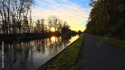 Rijkevorsel, Antwerpse Kempen, Belgium, sunset over canal path with trees soft golden light reflecting on calm water, bare tree silhouettes, empty asphalt path, crisp