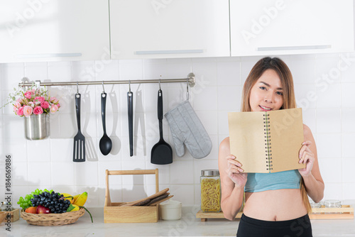 Smiling young asian woman in blue activewear hold up a blank notebook or recipe book in a modern kitchen, inviting idea for healthy cooking.