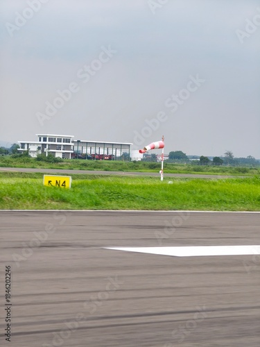 Red and white striped windsock blowing in the wind near a runway and airport building.