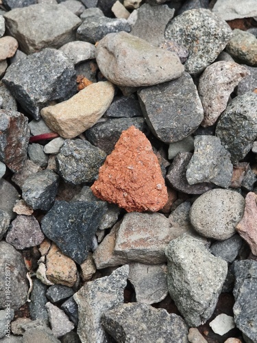 A triangular fragment of red brick lying among grey river stones and gravel rocks.