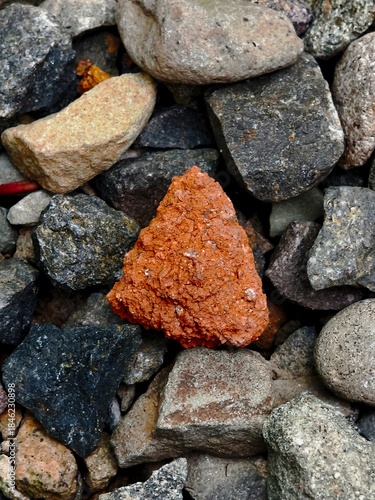 A triangular fragment of red brick lying among grey river stones and gravel rocks.