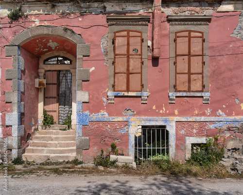 Facade of a dilapidated old house with crumbling pink plaster, rusty metal shutters, and a weathered stone archway. The abandoned building is overgrown with weeds