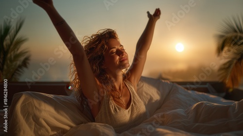 A beautiful young woman with curly hair stretches her arms with a happy smile while waking up in a comfortable white bed at sunrise, enjoying the warm morning light and a fresh start to the day.
