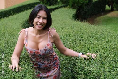 Beautiful young woman in a low-cut floral sundress smiles while leaning forward over a green garden hedge