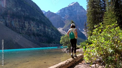 Young woman tourist walks carefully on a log over the turquoise water of the iconic moraine lake in banff national park, alberta, canada, enjoying the stunning view of the valley of the ten peaks