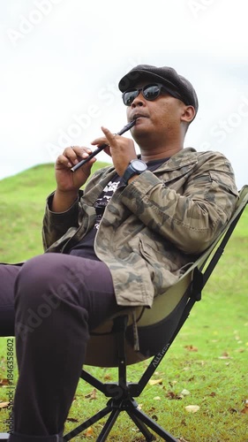 A male musician sits on a portable chair and play a tin whistle in a beautiful green meadow. A musician is playing a tin whistle in a green meadow with green hills in the background during the day