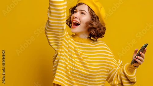 Happy woman celebrating success while holding a smartphone on yellow background