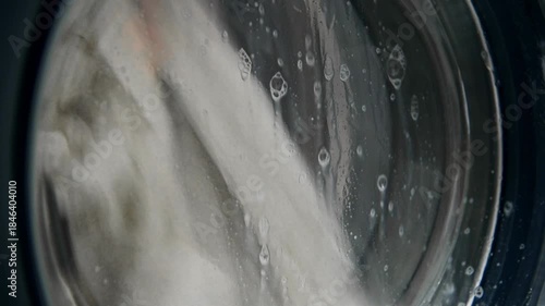 A close-up of a domestic washing machine window. White linens are washed, spinning and tumbling in the washing machine drum. Foam and bubbles run down the glass door viewing window.