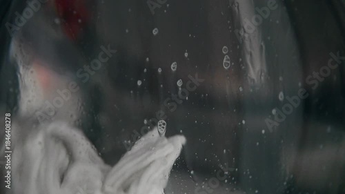 A close-up of a domestic washing machine window. White linens are washed, spinning and tumbling in the washing machine drum. Foam and bubbles run down the glass door viewing window.