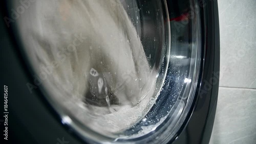 A close-up of a domestic washing machine window. White linens are washed, spinning and tumbling in the washing machine drum. Foam and bubbles run down the glass door viewing window.