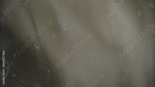 A close-up of a domestic washing machine window. White linens are washed, spinning and tumbling in the washing machine drum. Foam and bubbles run down the glass door viewing window.