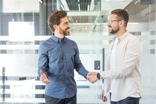 Φωτογραφία Two smiling adult businessmen standing in a modern office, shaking hands and dis