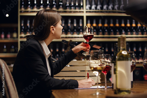 Fotografie Experienced female sommelier tasting wine into glass sitting in cellar over plenty bottle shelf background