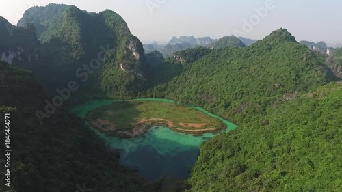 Aerial view of Trang An Valley in Vietnam: A vibrant turquoise lake is nestled between majestic, lushly forested limestone cliffs under a clear, sunny sky, showcasing iconic karst scenery.