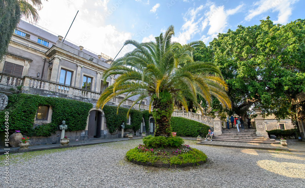 Fototapeta premium CATANIA, ITALY. The lush courtyard of Villa Cerami, a historic 18th-century palace currently housing the Faculty of Law, featuring a large palm tree, stone statues, and a grand stairc