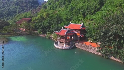 Aerial view of a picturesque small temple with a vibrant red roof on the shore of a turquoise lake. The building sits nestled against a forested cliffside in an idyllic, quiet bay.