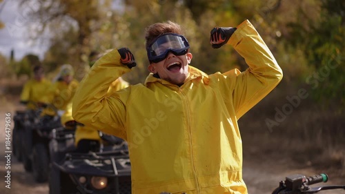 Slow motion. Group of white riders celebrating on quad trail, joyful leader pumps fists while friends in yellow jackets follow in line, dusty sunlit path and upbeat team