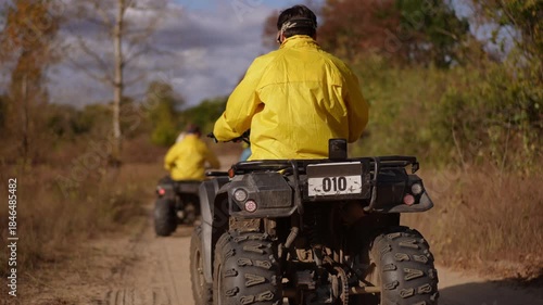 Slow motion. Rear view two atvs on trail close tandem ride with riders navigating rutted dirt track passing slender trees warm autumn light emphasis on companionship