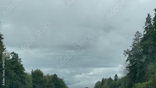 while driving in rain, raindrops remain on the windshield. A cloudy sky and trees on both sides frame the forward view.