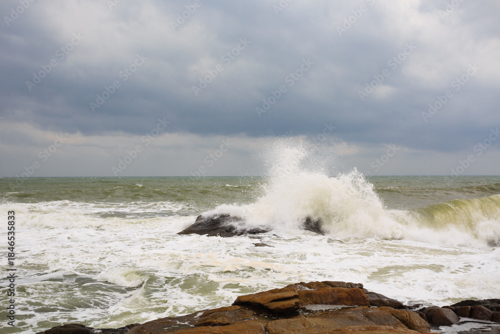 Fototapeta premium Under a cloudy sky, the waves crashed against the massive rocks, creating huge surges. (Scenic view of Hainan Stone Park, China)